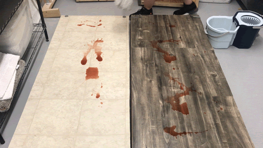 A mop moves across a section of linoleum flooring, mopping spilled red juice. Next to it, more spilled juice sits on a section of wood flooring.