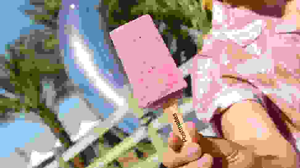 A person in a pink floral shirt holds a pink popsicle close to the camera lens.