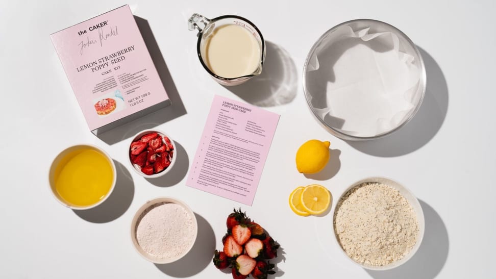 A topdown photo of The Caker's Lemon Strawberry Poppyseed pink boxed cake mix surrounded by the various ingredients that go into the cake—melted butter, freeze-dried strawberries, fresh strawberries, lemon juice, cake mix, and more—all on a white background.