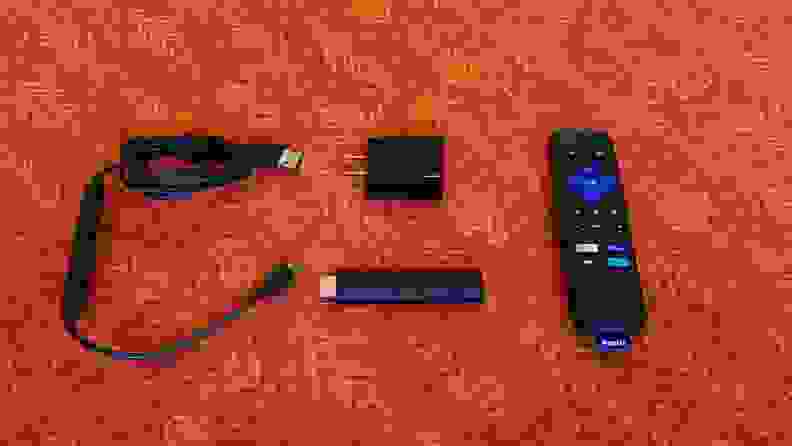 The matte black, candy-bar sized Roku Streaming stick is held with its port showing above a orange-red table with accessories.