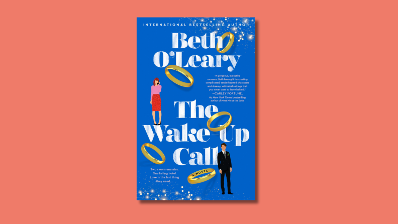 An image of the cover of 'The Wake-Up Call' by Beth O'Leary featuring a couple in business clothes surrounded by falling gold rings.
