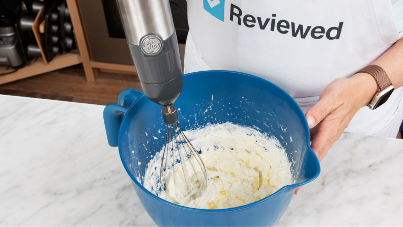 Person with apron on stands at kitchen countertop while using GE Immersion Blender with whisk attachment to whip cream inside of blue bowl.