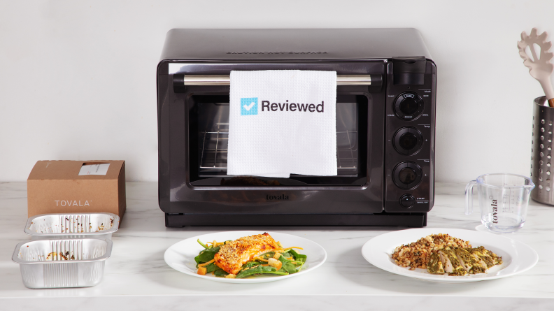 The Tovala Smart Oven Pro in a white kitchen with two plates of prepared food in front of it and a Reviewed towel hanging from the handle.