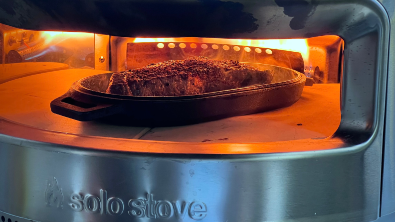 Small steak cooking inside of Solo Stove Pizza Oven on cast iron dish outdoors.